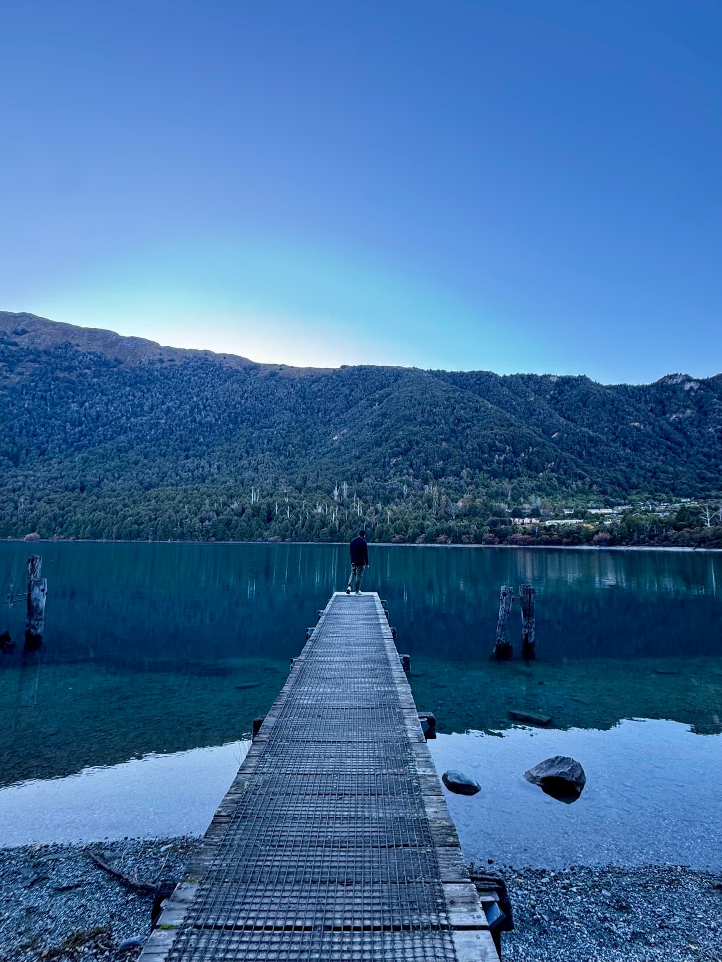 A lone figure standing at the end of a jetty, still lake and mountains ahead